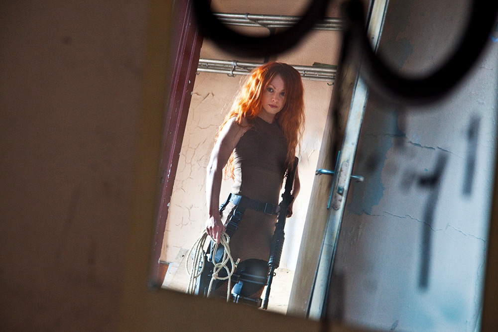 Spegelbild på kvinna med rött långt hår som står i dörröppning med rep i ena handen och ett maskingevär i den andra.