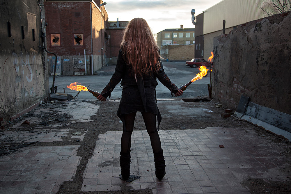 En svartklädd kvinna med långt rött hår, bakifrån, håller två brinnande molotovcocktails i vardera hand