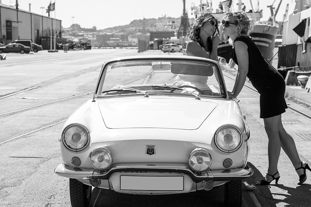 En kvinna i klänning och solglasögon sitter i en vit fransk gammal Renault Cabriolet och pratar med en kvinna i svart klänning och solglasögon som lutar sig mot bilen.
