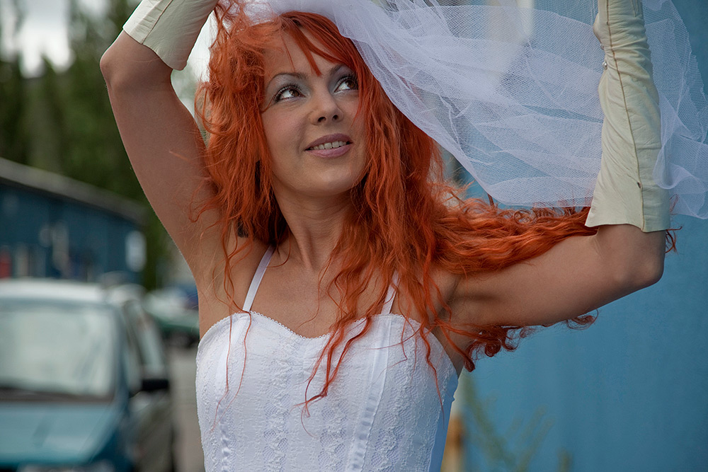 Leende kvinna med långt rött hår i bröllopsklänning och långa vita handskar höjer armarna ovanför huvudet och rättar till sitt flor
